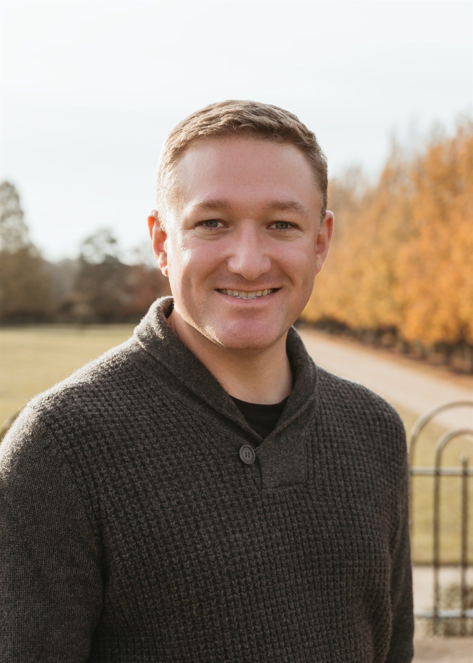 Max Triantos smiling outdoors in a dark sweater, autumn trees behind him.